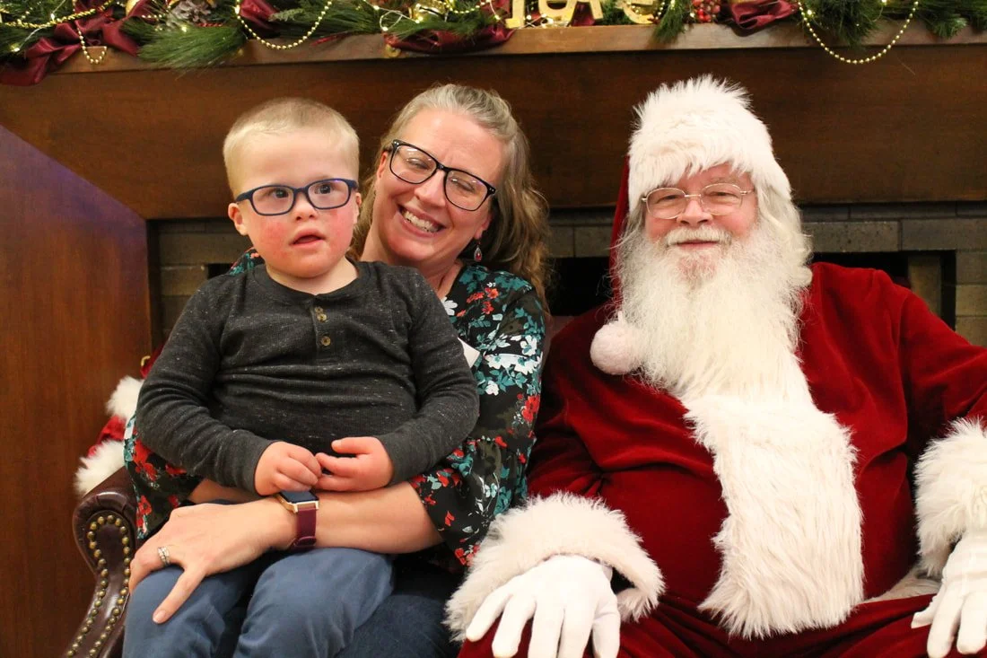 A woman, a young boy, and Santa Claus sitting together on a couch in front of a decorated fireplace mantel. The woman is smiling, the boy looks surprised, and Santa is smiling with his eyes closed.
