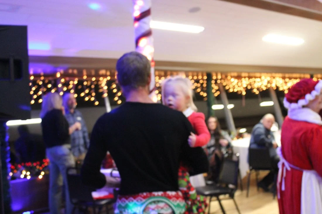 A man holding a young girl in a festive holiday party with Christmas lights and people in the background.