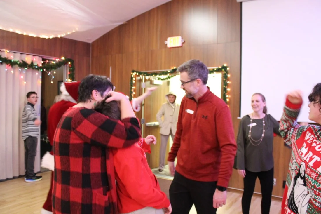 People celebrating at a Christmas holiday party, some wearing festive clothing like Santa hats and Christmas sweaters, in a decorated room with string lights and holiday decorations.