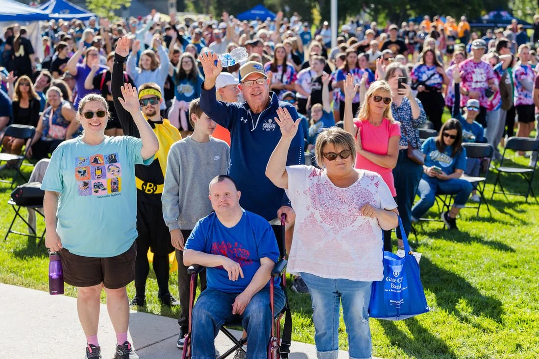 A large group of people gathered outdoors, some of them are raising their hands in a wave or greeting, during a sunny day at an event or gathering.