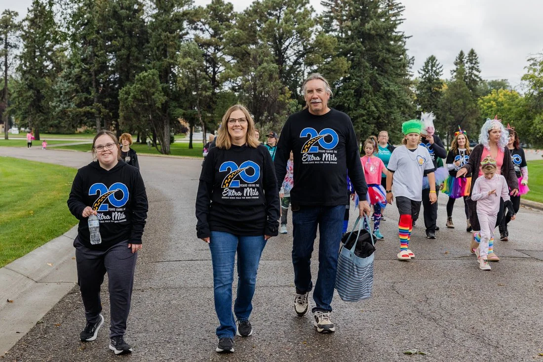 People participating in a walk or parade, some wearing matching T-shirts celebrating a 20th anniversary of something, with children in colorful costumes and unicorn hats, walking on a park pathway.