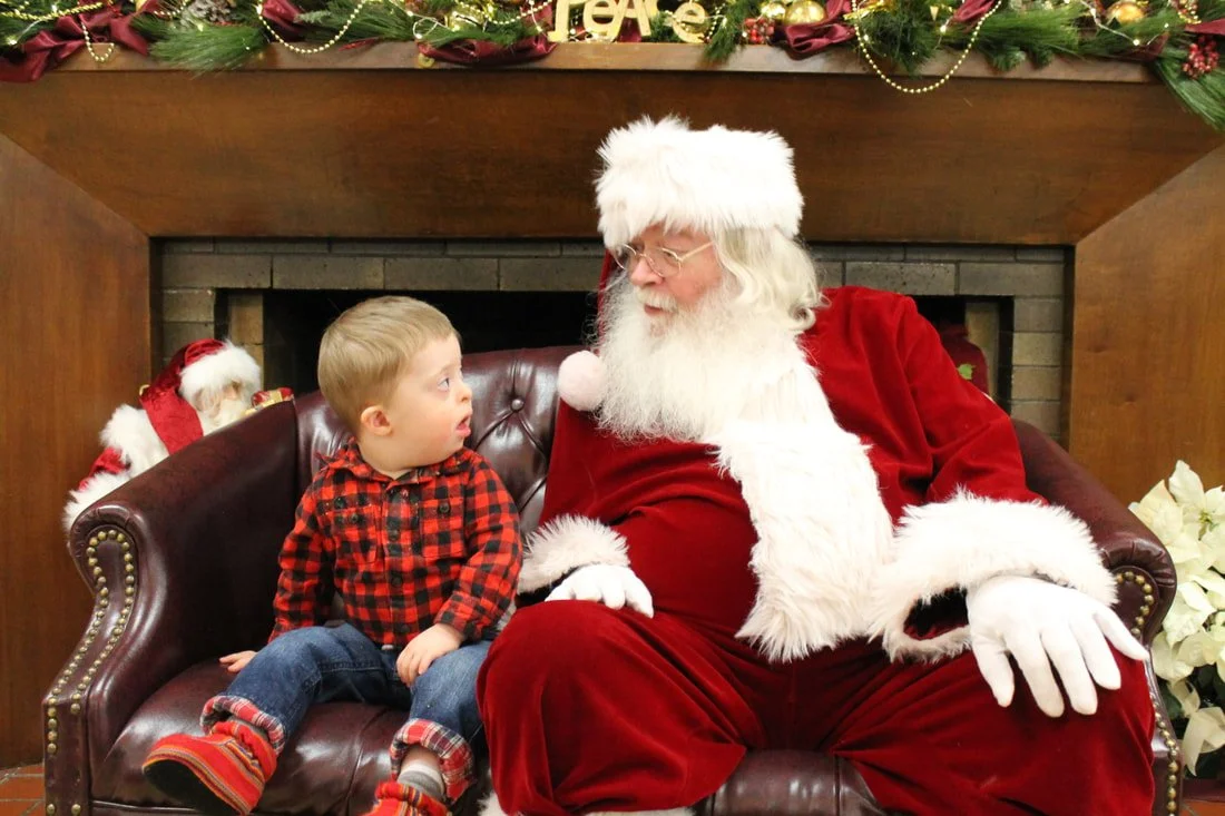 A young boy in a red plaid shirt sitting on a leather couch next to Santa Claus, who is dressed in a red suit with white fur trim and glasses, inside a room decorated for Christmas with holiday garland, and a fireplace in the background.