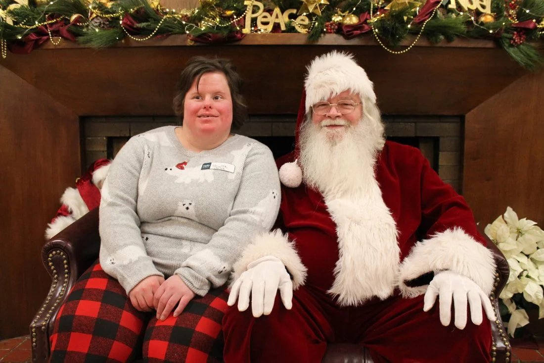 A woman with short brown hair and a gray sweater sitting next to a man dressed as Santa Claus with a white beard, red suit, white gloves, and glasses, sitting in front of a decorated fireplace with holiday garlands, ornaments, and poinsettias.
