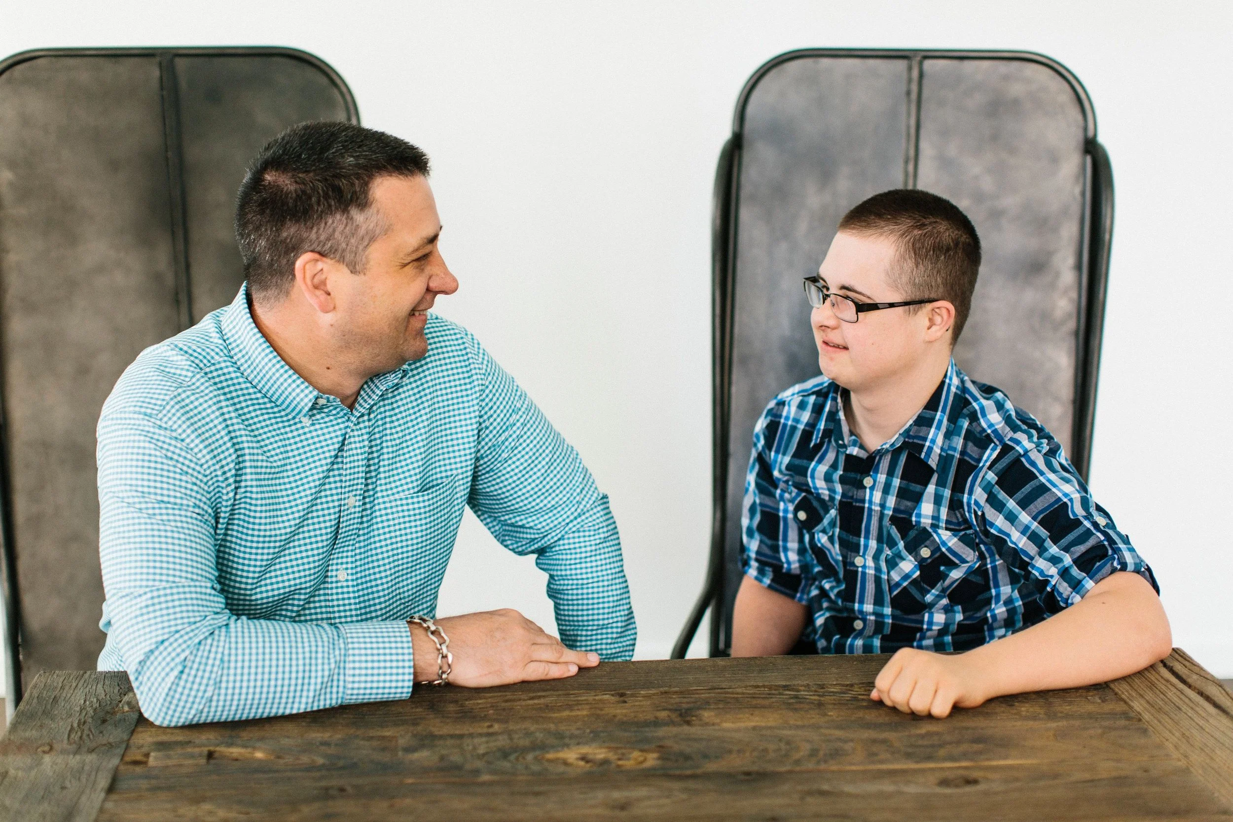 An adult man and a boy sitting across from each other at a wooden table, smiling and engaging in a conversation, with grey chairs behind them.