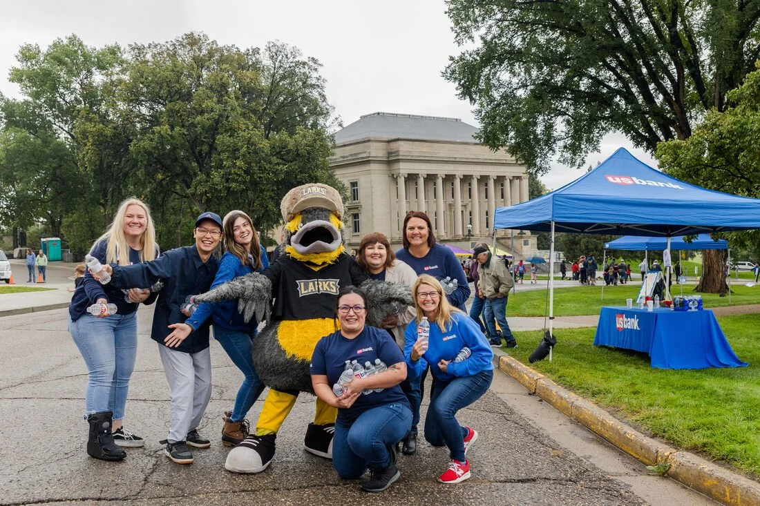 Group of people, including a person in a green and yellow lizard mascot costume, smiling and posing for a photo outdoors in front of a government building with trees and a blue USPS bank tent nearby, on a cloudy day.