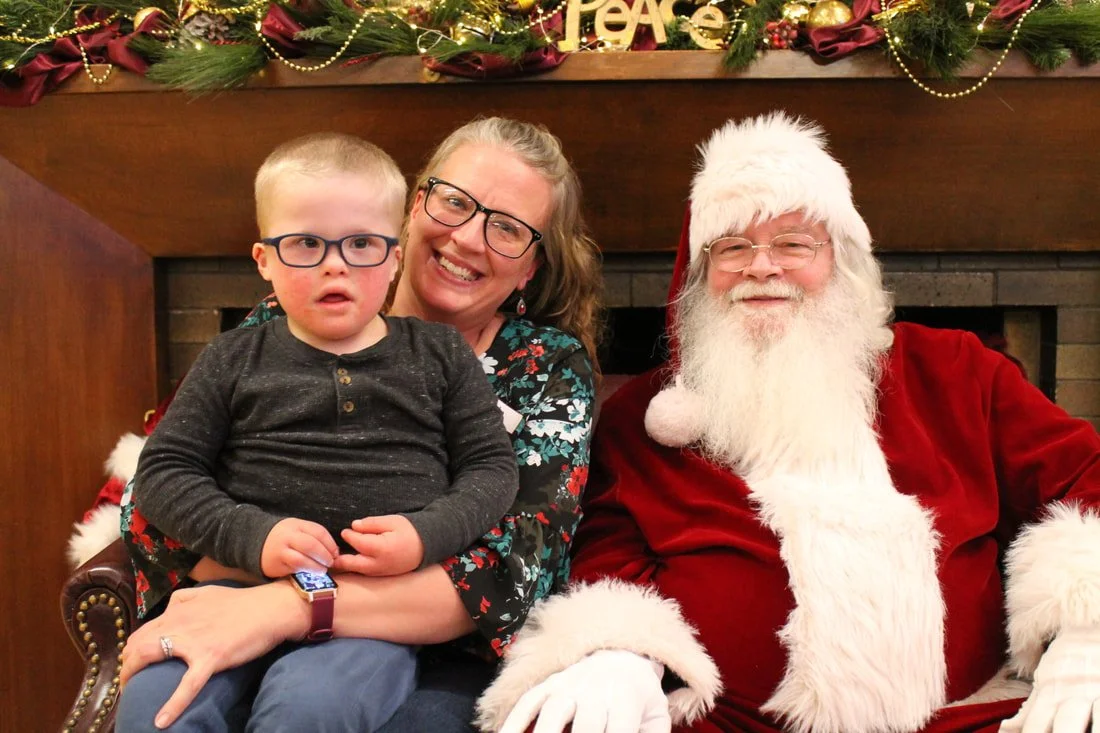A woman with glasses and a child sitting on her lap, smiling, next to Santa Claus at a Christmas celebration. The background features holiday decorations and a fireplace.