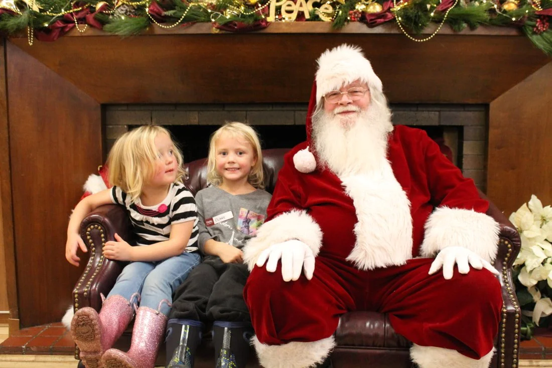 Two young girls sitting on Santa's lap, posing for a photo in front of a decorated fireplace with garlands and poinsettias, during a Christmas celebration.