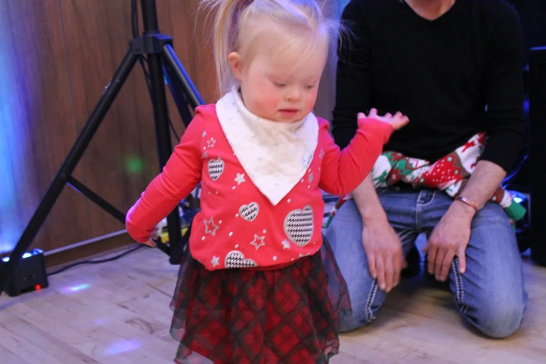 A young girl with blonde hair wearing a red sweater with black and white heart and star patterns, a white bib, and a red plaid skirt dancing or moving with her eyes closed, while an adult in a black shirt and blue jeans with Christmas-themed pajamas 