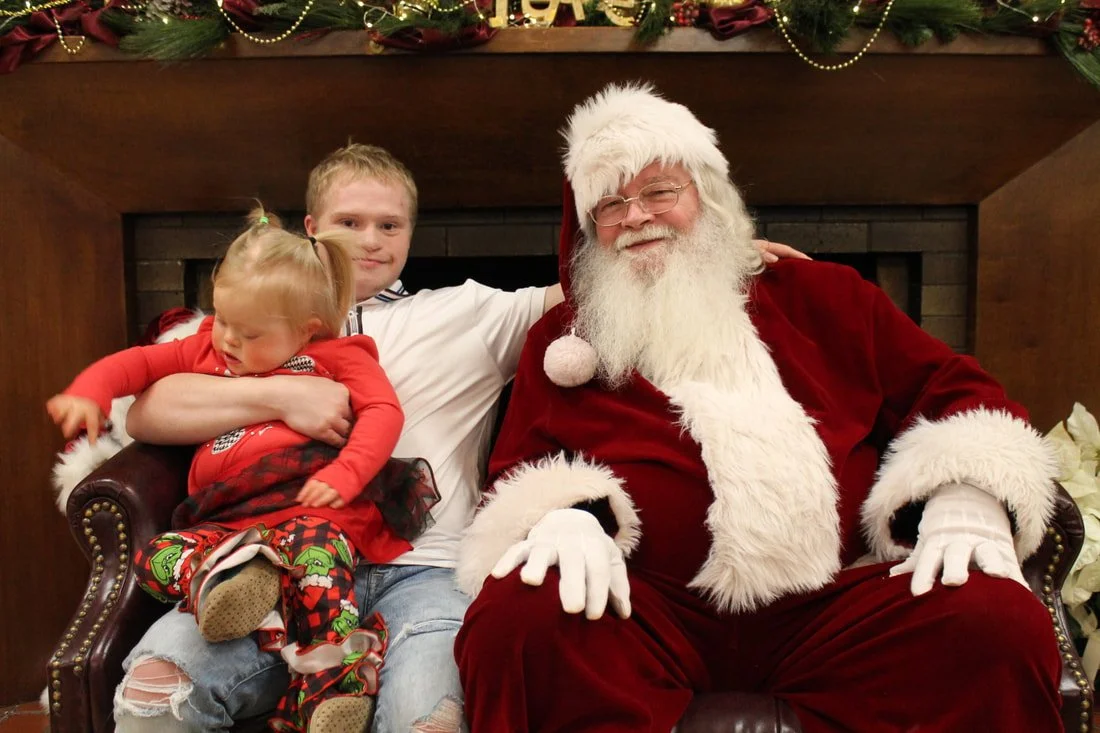 A family sitting with Santa Claus near a fireplace decorated for Christmas, with a young girl sitting on the arm of the man, and Santa smiling