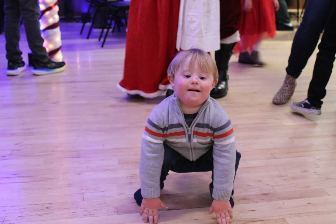 A young boy kneeling on the wooden floor at a Christmas party, surrounded by people in festive attire.