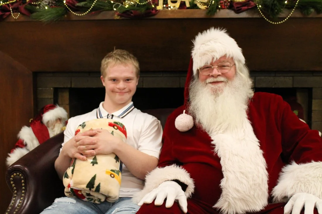 A boy sitting with Santa Claus in front of a fireplace, holding a Christmas present, with Christmas decorations above.