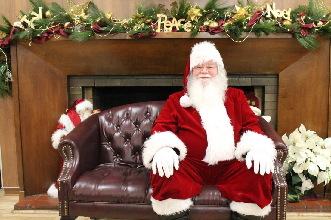 A man dressed as Santa Claus sitting on a leather couch in front of a decorated fireplace with Christmas garland, ornaments, and words like 'Noel,' 'Peace,' and 'Joy' hanging on the mantel. There are poinsettia plants on each side of the fireplace.