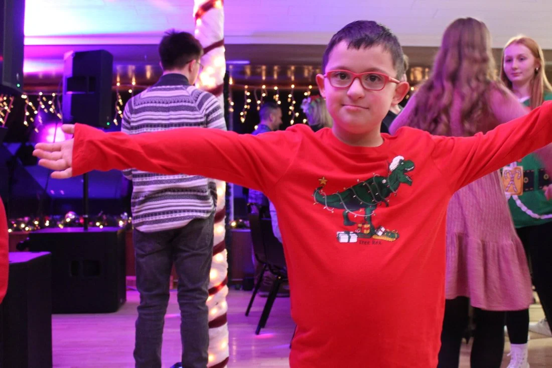 A young boy with glasses and a Christmas-themed shirt with a dinosaur on it, standing with arms outstretched at a holiday party with festive lights and other children in the background.