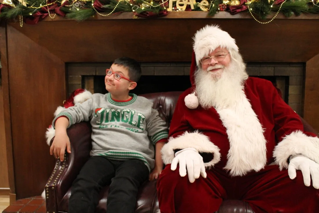 A young boy sitting next to Santa Claus, both smiling, in front of a fireplace decorated with Christmas garland and ornaments.