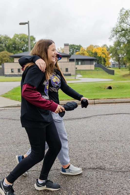 Two young girls are outdoors, one is hugging the other from behind while they walk on a sidewalk. The girl in front is smiling and holding a camera in her right hand, wearing a black and maroon hoodie, black leggings, and sneakers. The girl behind is