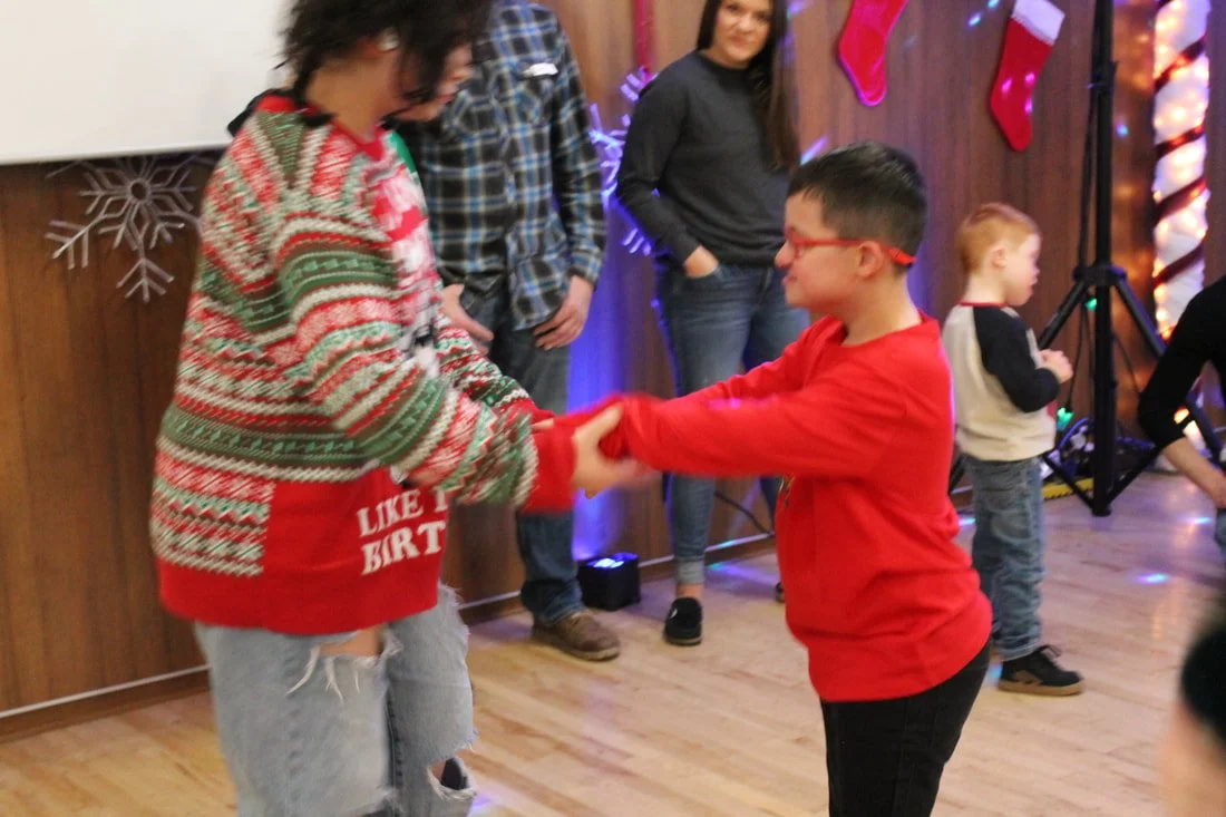 A boy and a woman are dancing together at a Christmas party, holding hands and smiling. The woman is wearing a festive sweater, and the boy is dressed in a red shirt. In the background, there are Christmas decorations and other children and adults wa