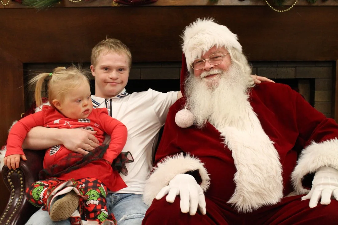 A young girl with blonde hair in pigtails sitting on a man's lap, a young boy sitting next to her, and Santa Claus dressed in a red suit with white fur trim, wearing glasses, sitting in front of a fireplace.