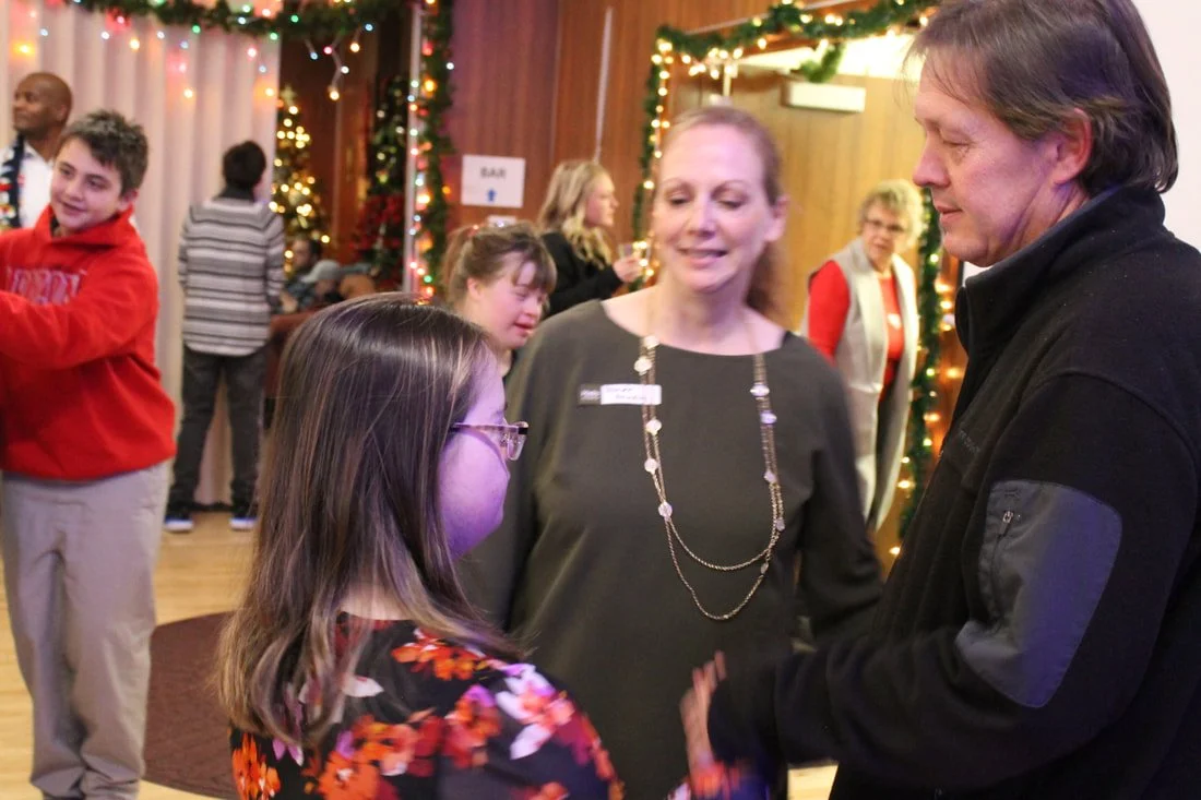 A group of people at a holiday gathering, with Christmas lights and decorations in the background. A young girl with glasses and a floral top is speaking with a man, while a woman stands nearby listening.
