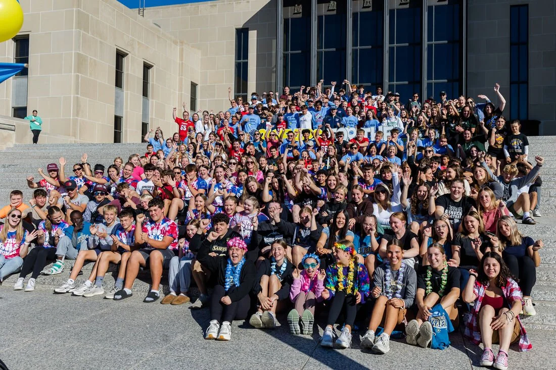 Large group of young students gather on steps outside a building, posing for a group photo, wearing casual clothes and some with sunglasses, hats, and accessories.