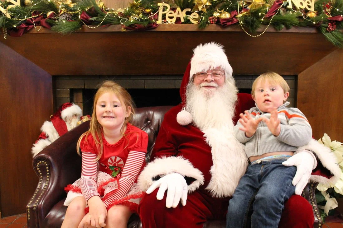 Santa Claus sitting on a leather couch with two children, a young girl and a young boy, in a festive Christmas setting with decorations, garland, and holiday words above them.
