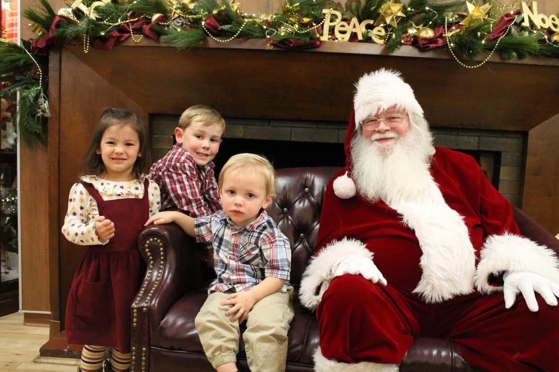 Three young children sitting with Santa Claus in front of a decorated fireplace with festive Christmas decorations, including greenery, gold ornaments, and 'Peace' and 'Noel' signs.