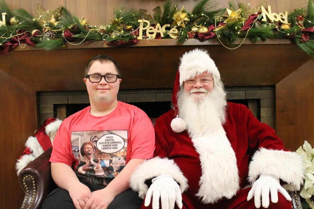 A young man sitting next to Santa Claus, both smiling, in front of a decorated fireplace with Christmas garlands, ornaments, and signs reading 'Peace', 'Noel', and 'Xmas'.