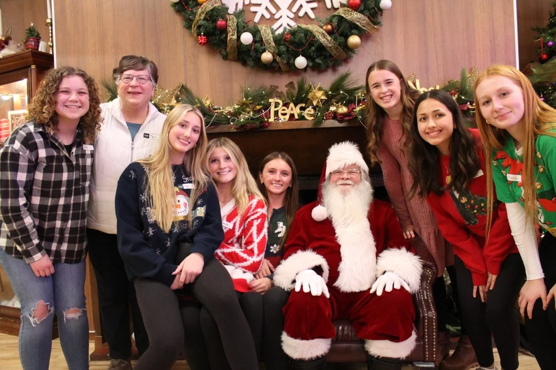 Group of smiling children and a woman with Santa Claus at a Christmas event, with Christmas decorations including a wreath and garland in the background.