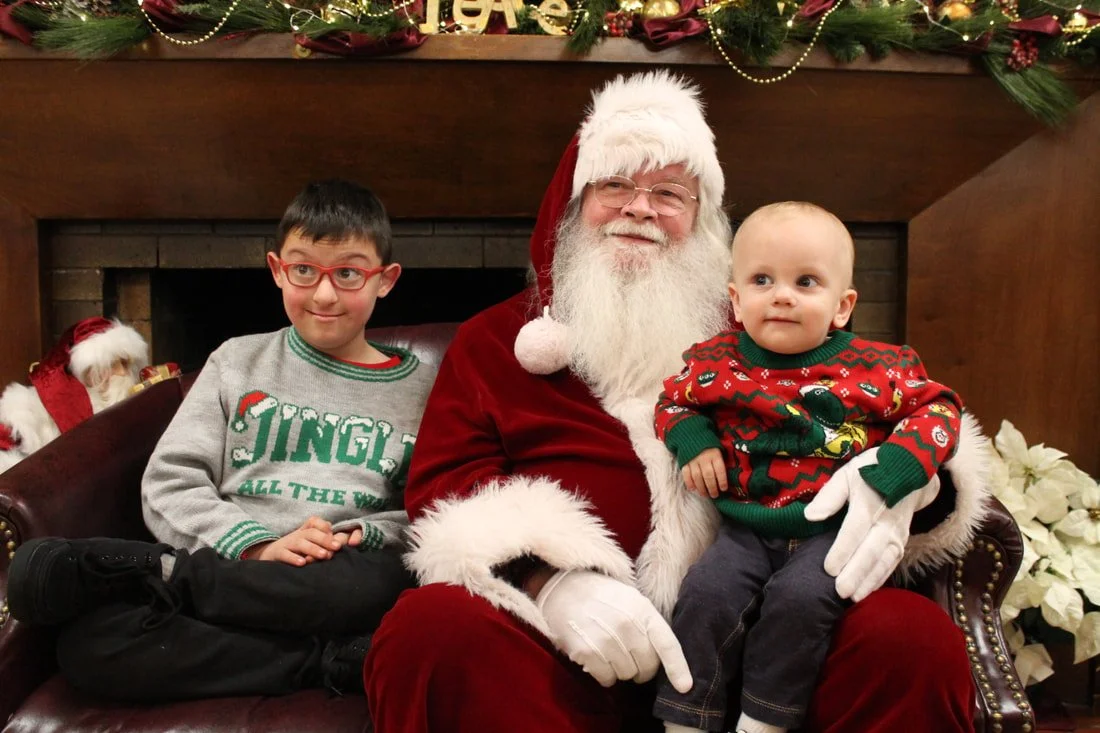 A man dressed as Santa Claus sitting on a red chair with two children, one older boy with glasses and a Christmas sweater, and a younger child in a festive sweater, all in front of a holiday mantle decorated with ornaments and greenery.