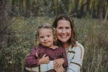 A woman and a young girl smiling outdoors in a wooded area, with the woman holding the girl in her arms.