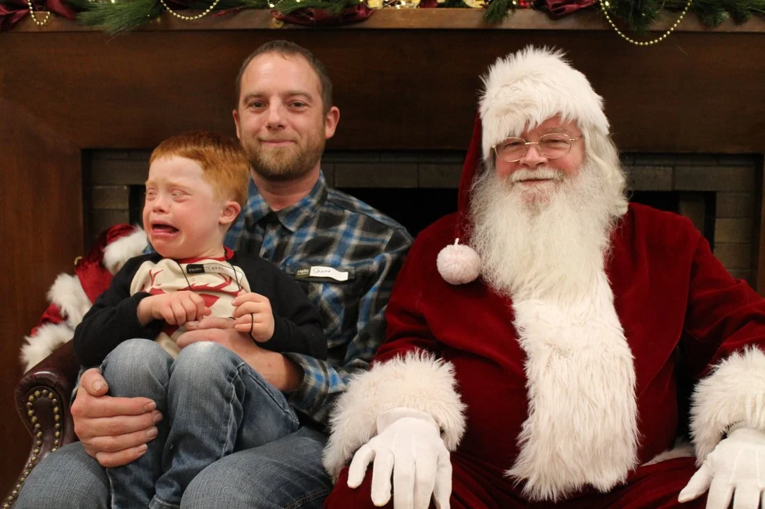 A young boy crying and sitting on a man's lap next to Santa Claus, who is smiling. They are in front of a fireplace decorated for Christmas.