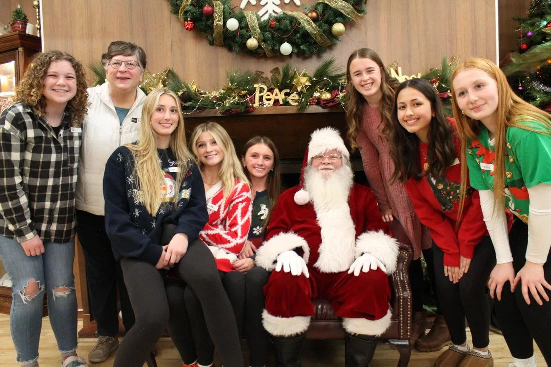 Group of eight girls and a woman with short gray hair posing with Santa Claus, who is seated, in front of a decorated Christmas tree and a wreath.
