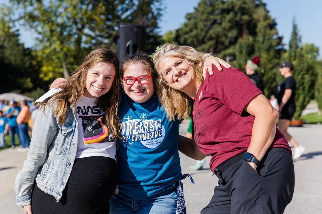 Three smiling women with their arms around each other, outdoors at a sunny event with a crowd and trees in the background.