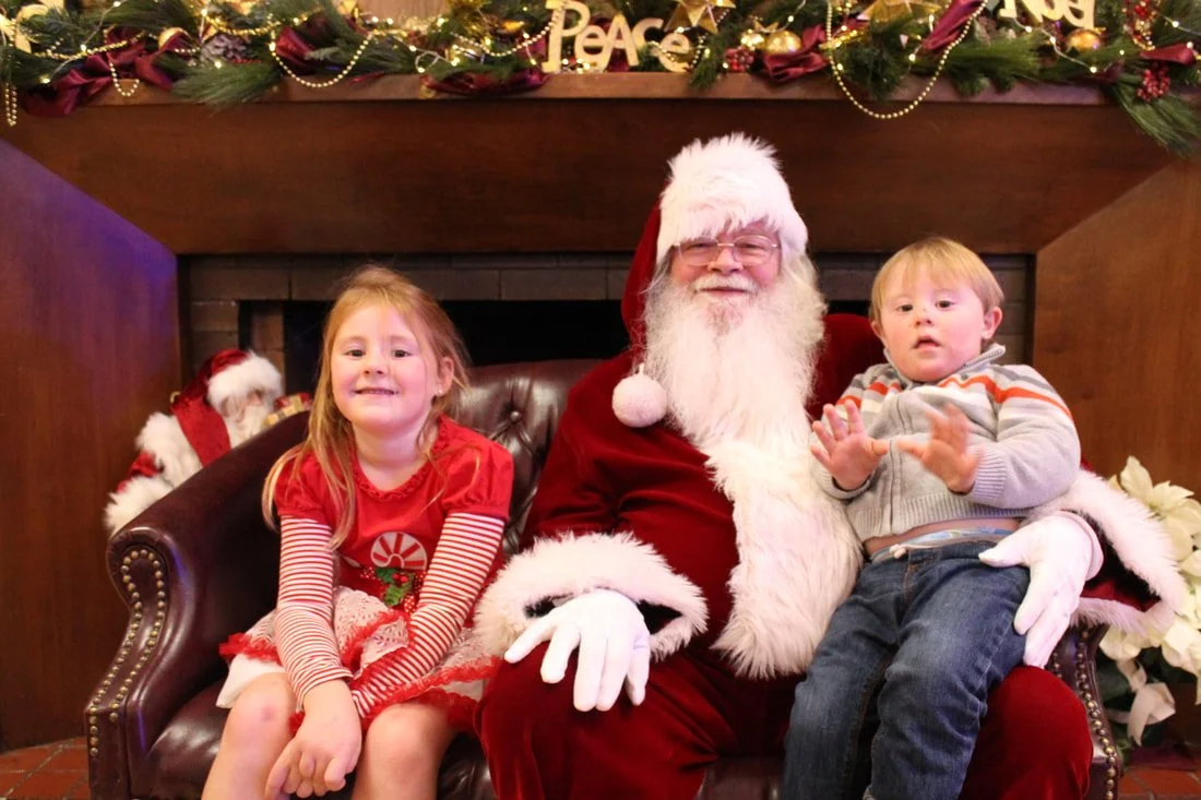 A young girl and a young boy sitting with Santa Claus for a Christmas photo. The girl is wearing a red dress with striped sleeves and red gloves, smiling. The boy, wearing a gray sweater with orange and white stripes, is sitting on Santa's lap, looki