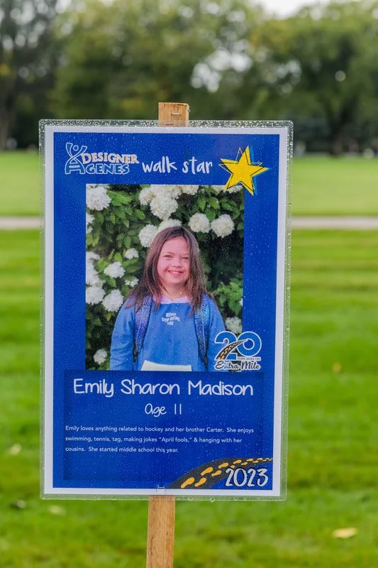 Outdoor sign featuring a young girl named Emily Sharon Madison, age 11, with a big smile, standing in front of white flowering bushes. The sign is blue with yellow and white accents, displaying text about her interests and achievements, including a 2