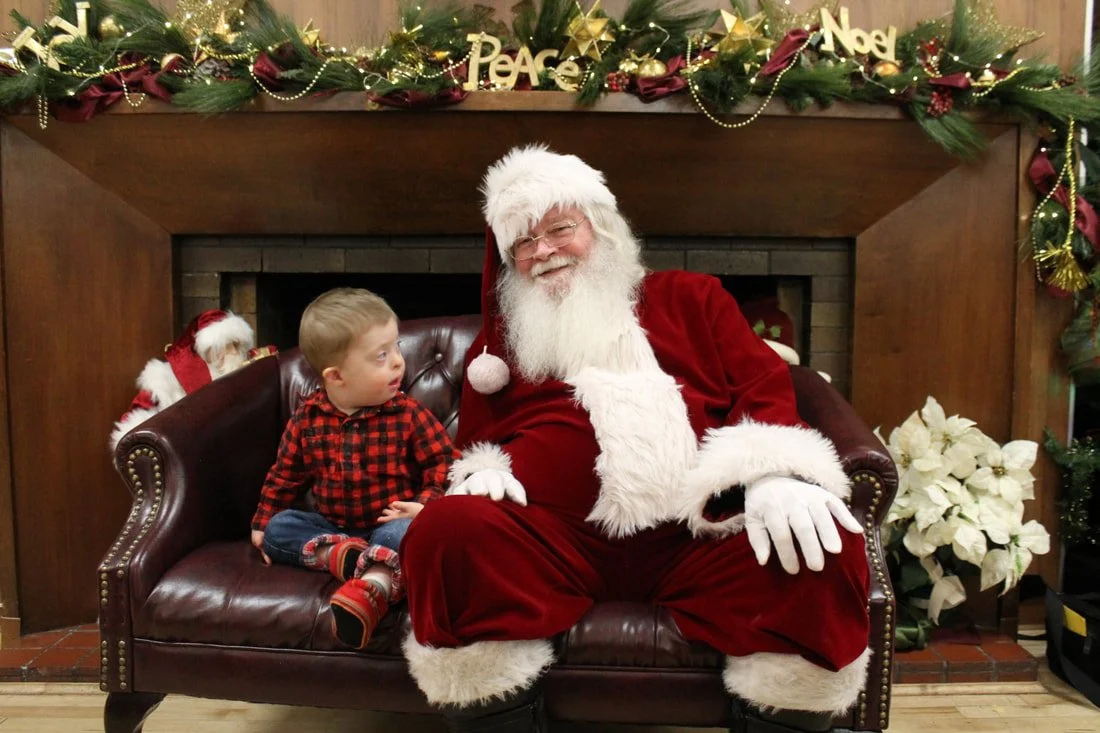 A young boy in a red and black checkered shirt sitting on a dark brown leather couch next to a smiling man dressed as Santa Claus in a red suit with white fur trim. They are in front of a fireplace decorated with Christmas garland, gold ornaments, an