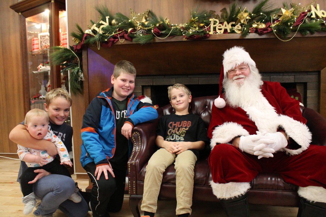 Four children sitting with Santa Claus on a leather sofa in front of a decorated fireplace with Christmas garland and ornaments.