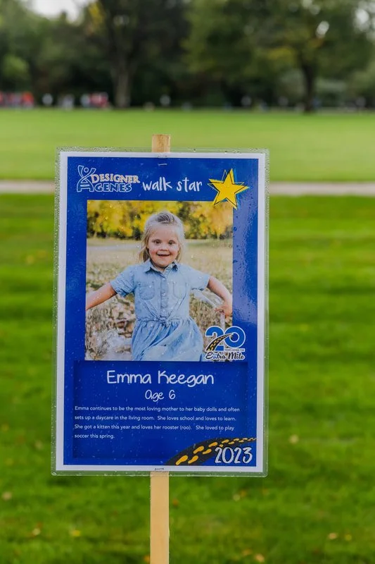 A sign featuring a young girl named Emma Keegan, age 6, smiling outdoors with trees in the background. The sign has a blue border with text, a star graphic, and a 20th anniversary logo for extra mile walking event, dated 2013.