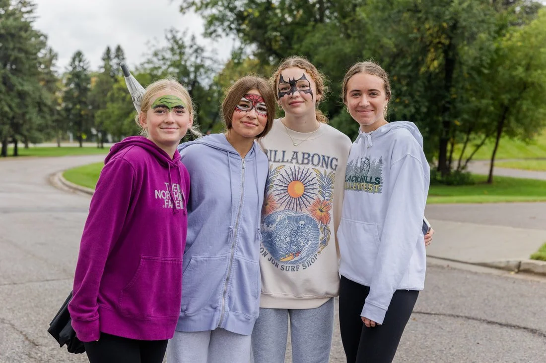 Four girls with face paint standing outdoors on a paved pathway with trees in the background.