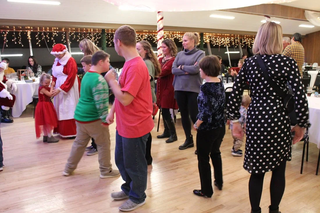 People gathered at a Christmas party, some standing and some dancing, with Christmas decorations and fairy lights in the background.
