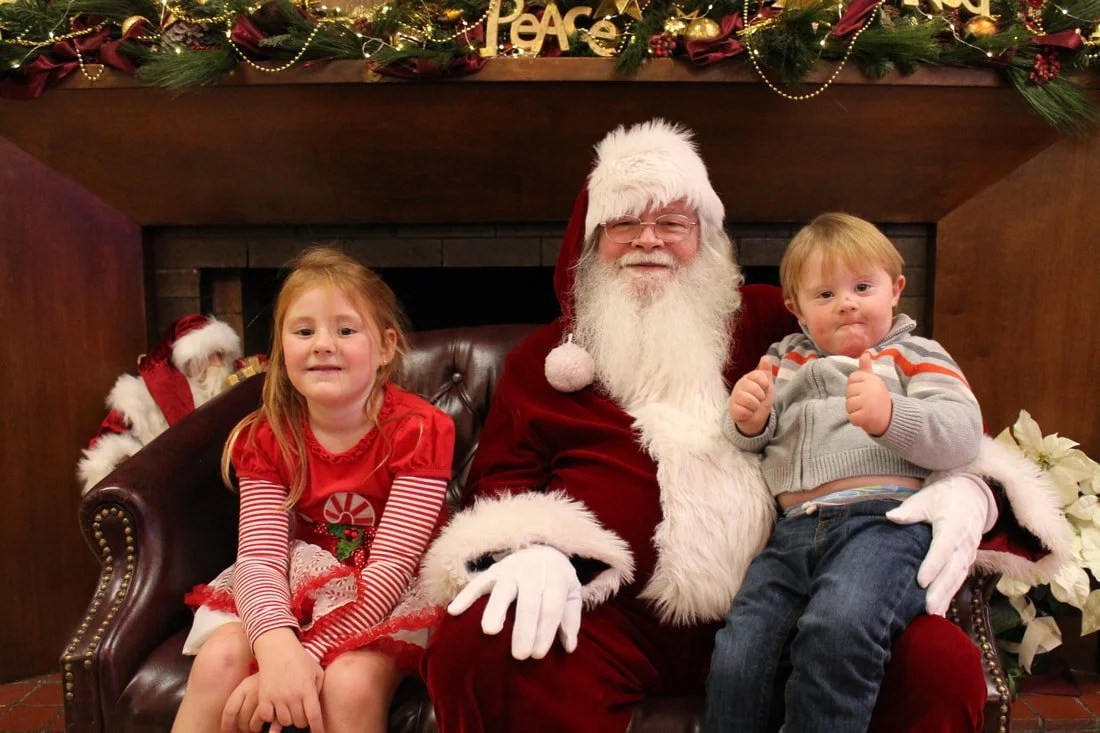 A man dressed as Santa Claus sitting on a bench with a young girl and a young boy, all smiling. The girl is wearing a red holiday dress with striped sleeves, and the boy is wearing a gray sweater with orange and white stripes. They are in front of a 