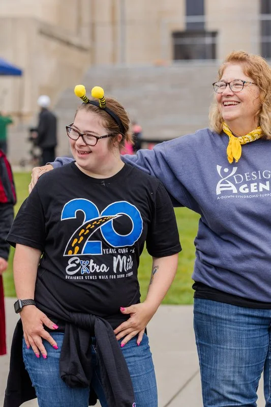Two women, one young girl and one older woman, are smiling and standing outdoors. The young girl is wearing a black t-shirt with a 20th anniversary design and yellow headband. The older woman is wearing glasses, a blue sweatshirt with a logo, and a y