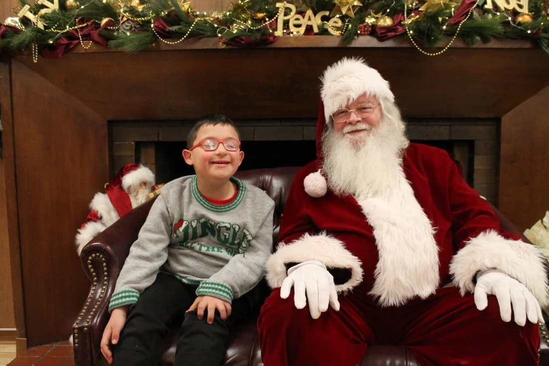 A young boy with red glasses sitting on Santa's lap, both smiling, in a Christmas setting with Christmas decorations and the word 'Peace' above.