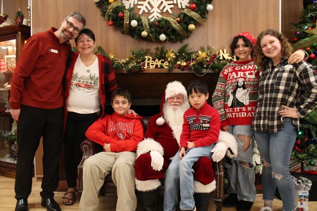 Group of people with Santa Claus during Christmas celebration, standing in front of a decorated fireplace and Christmas wreath.