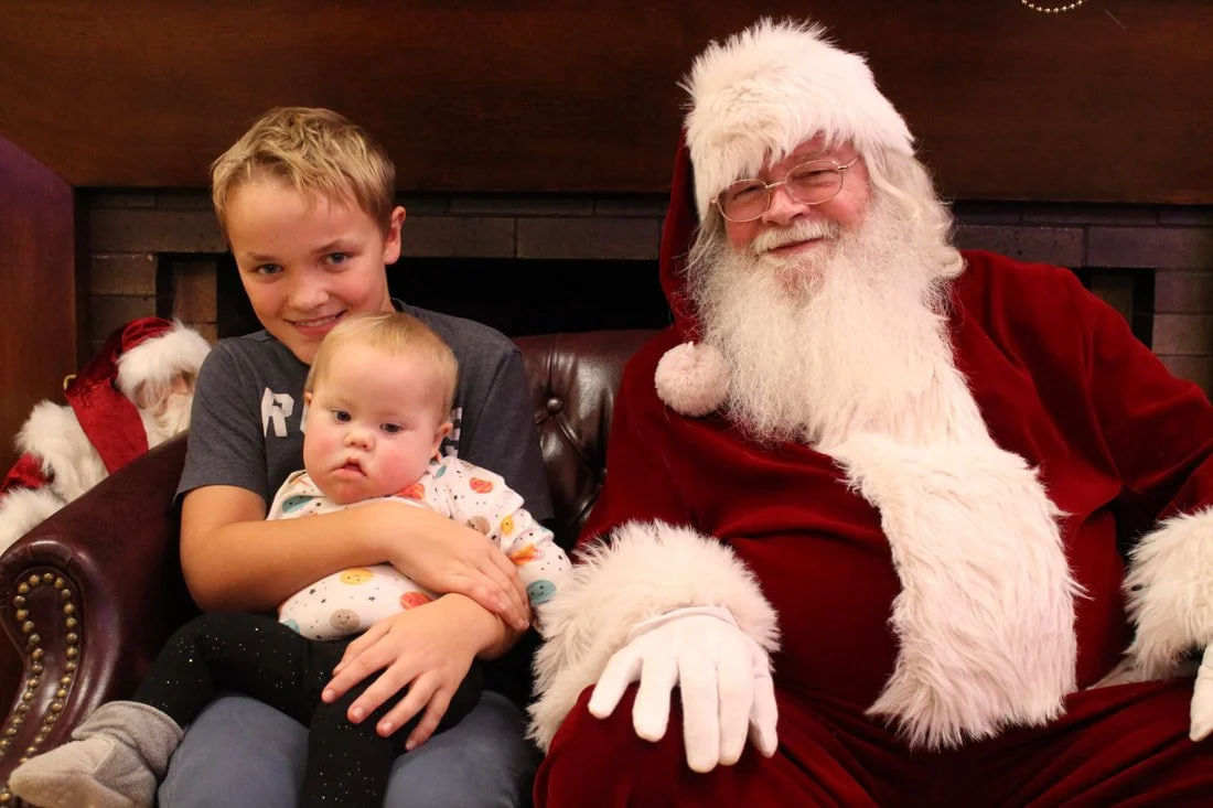 A young boy and a baby girl sitting on Santa Claus's lap in front of a fireplace.