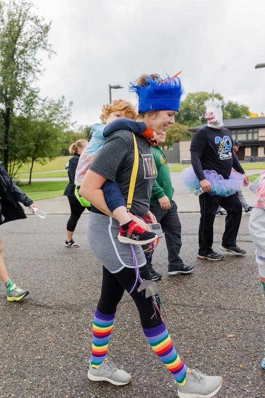 People participating in a festive outdoor event, including a woman with rainbow-striped knee-high socks, wearing a blue wig, and a person dressed in a unicorn costume.