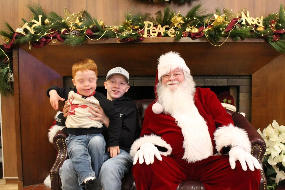 Two young boys sitting on Santa Claus's lap in front of a decorated fireplace with holiday garland, ornaments, and words like 'Peace' and 'Noel', celebrating Christmas.