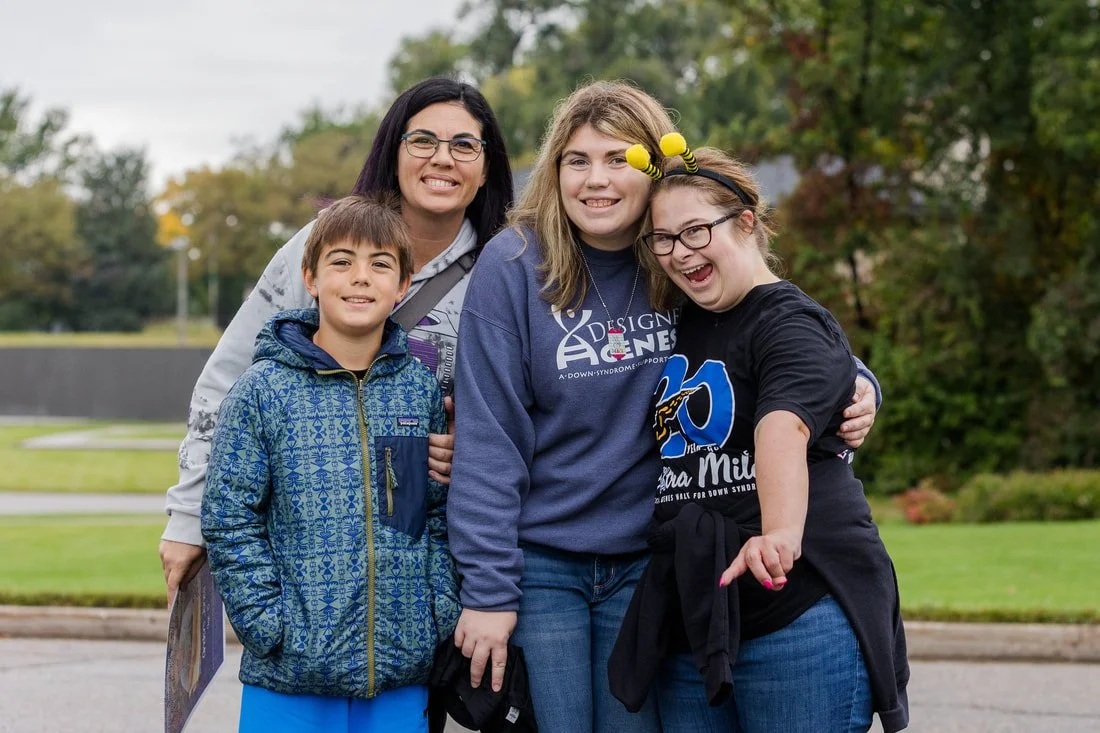 Group of four people, two adults and two children, smiling outdoors in a park with trees and grass.