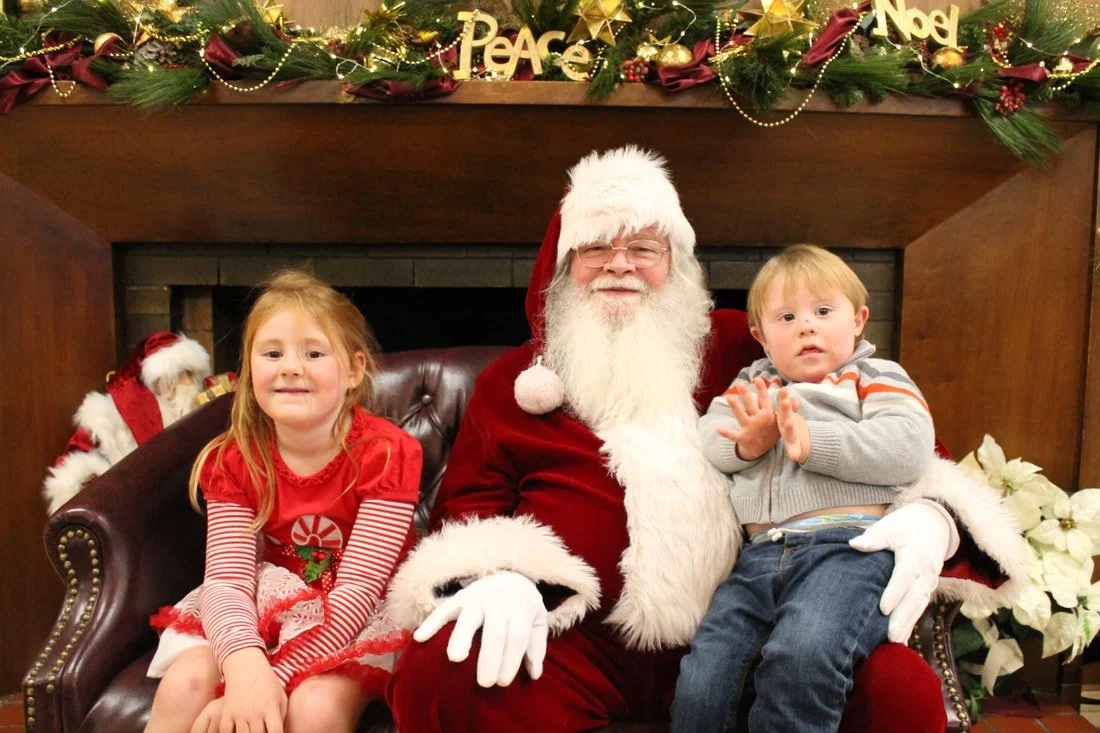 A man dressed as Santa Claus sitting on a leather couch flanked by two young children, a girl and a boy, in a festive Christmas setting with holiday decorations and flowers.