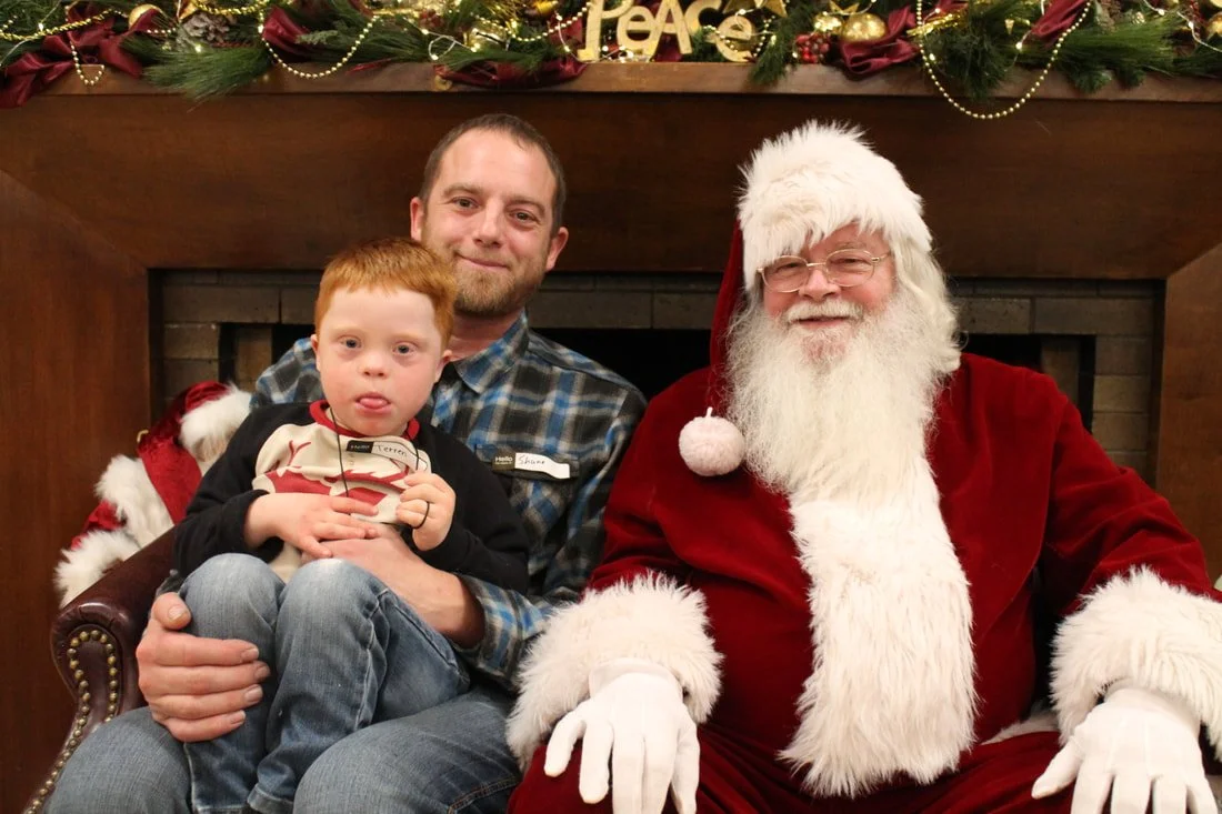 A man with a young boy sitting next to Santa Claus, who is dressed in a red suit with a white beard and hat, in front of a fireplace decorated with Christmas garlands, ornaments, and a 'Peace' sign.
