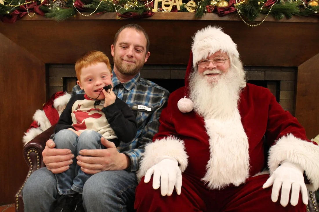 A young boy sitting on a man's lap next to Santa Claus, all smiling in front of a Christmas fireplace.
